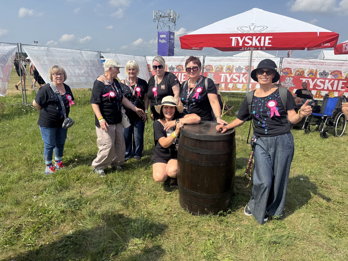 Siedem Mam zebrało się wokół drewnianej beczki w strefie Tyskiego. Jedna kuca przy beczce, druga pokazuje kciuk w górę, a na ich czarnych koszulkach widać różowe przypinki „Oddech dla MAM”.